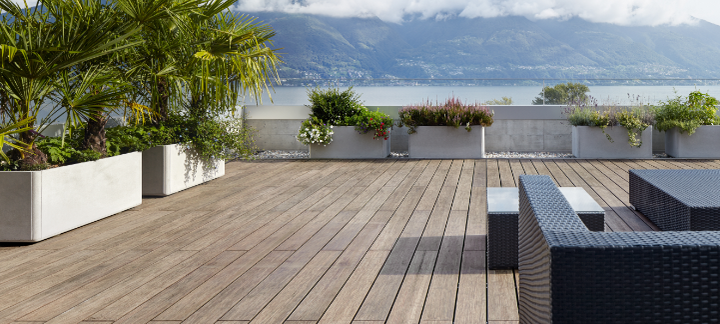 Große Terrasse mit Bambus-Holzdielen, modernen Rattanmöbeln, Pflanzen in Betonkübeln und Blick auf einen See mit Bergen.