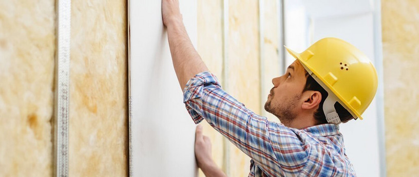 Handwerker mit gelbem Schutzhelm montiert eine Gipskartonplatte auf eine Metallständerwand mit Dämmung.