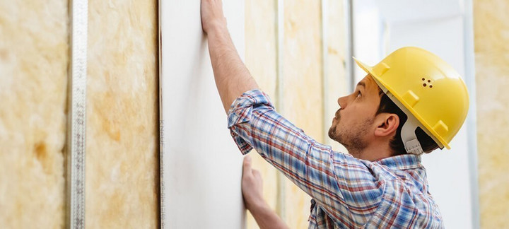 Handwerker mit gelbem Schutzhelm montiert eine Gipskartonplatte auf eine Metallständerwand mit Dämmung.