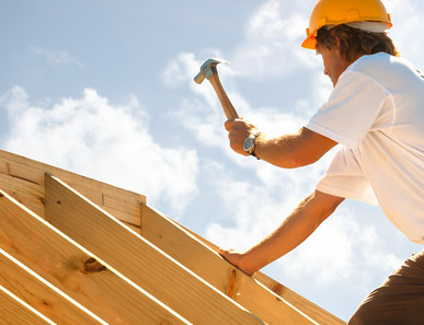Dachdecker mit gelbem Schutzhelm beim Befestigen von Konstruktionsvollholz (KVH) auf einem Dachstuhl unter strahlendem Himmel.