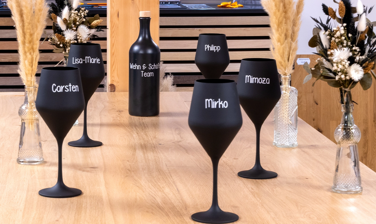 Schwarze Weingläser mit den Namen der Mitglieder des Wehn & Schäfer Teams beschriftet, dazu eine Flasche mit Aufschrift 'Wehn & Schäfer Team' auf einem Holztisch.
